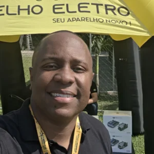 Anderson Coelho de camisa polo preta com a logo da Coelho Eletro, em frente à tenda amarela da loja em um evento