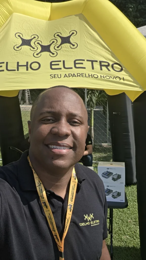 Anderson Coelho de camisa polo preta com a logo da Coelho Eletro, em frente à tenda amarela da loja em um evento