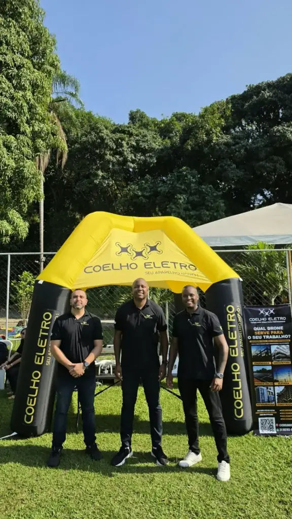 Três integrantes da equipe da Coelho Eletro de camisa polo preta em frente à tenda inflável amarela