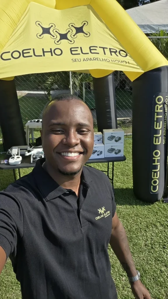 Integrante da equipe da Coelho Eletro sorrindo em frente à tenda amarela com drones na mesa